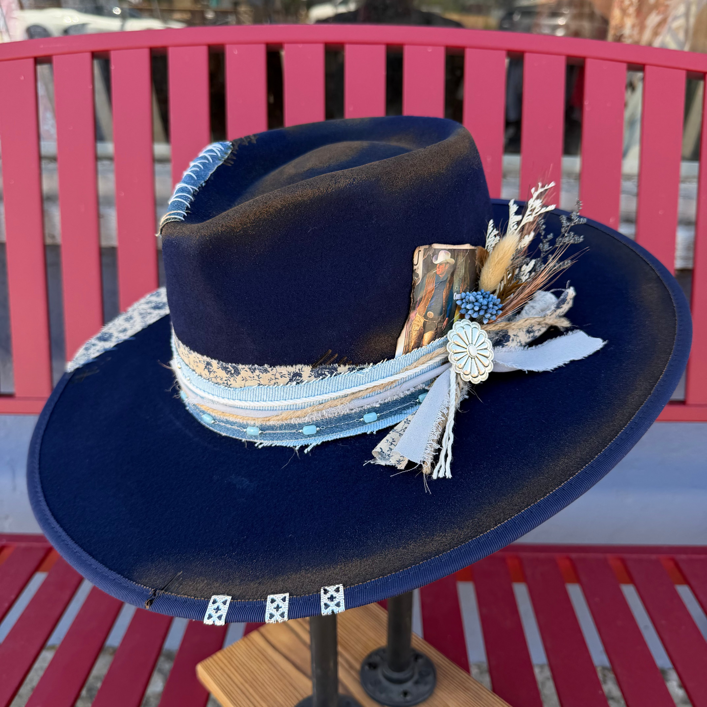 A navy blue wide-brim fedora hat displayed on a wooden stand, featuring a decorative band with light blue and beige stripes, dried grasses, and a small white flower embellishment, set against a red bench background.
